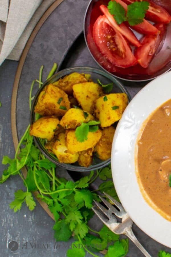Cumin-Spiced Potatoes in small bowl with butter chicken and sliced tomatoes